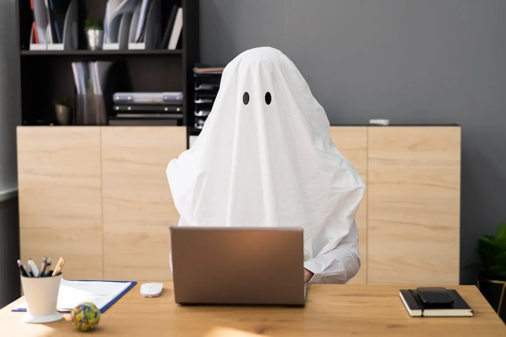 Person covered with a ghost sheet working on a laptop, symbolizing ghostwriting services for businesses at BigSEO.net